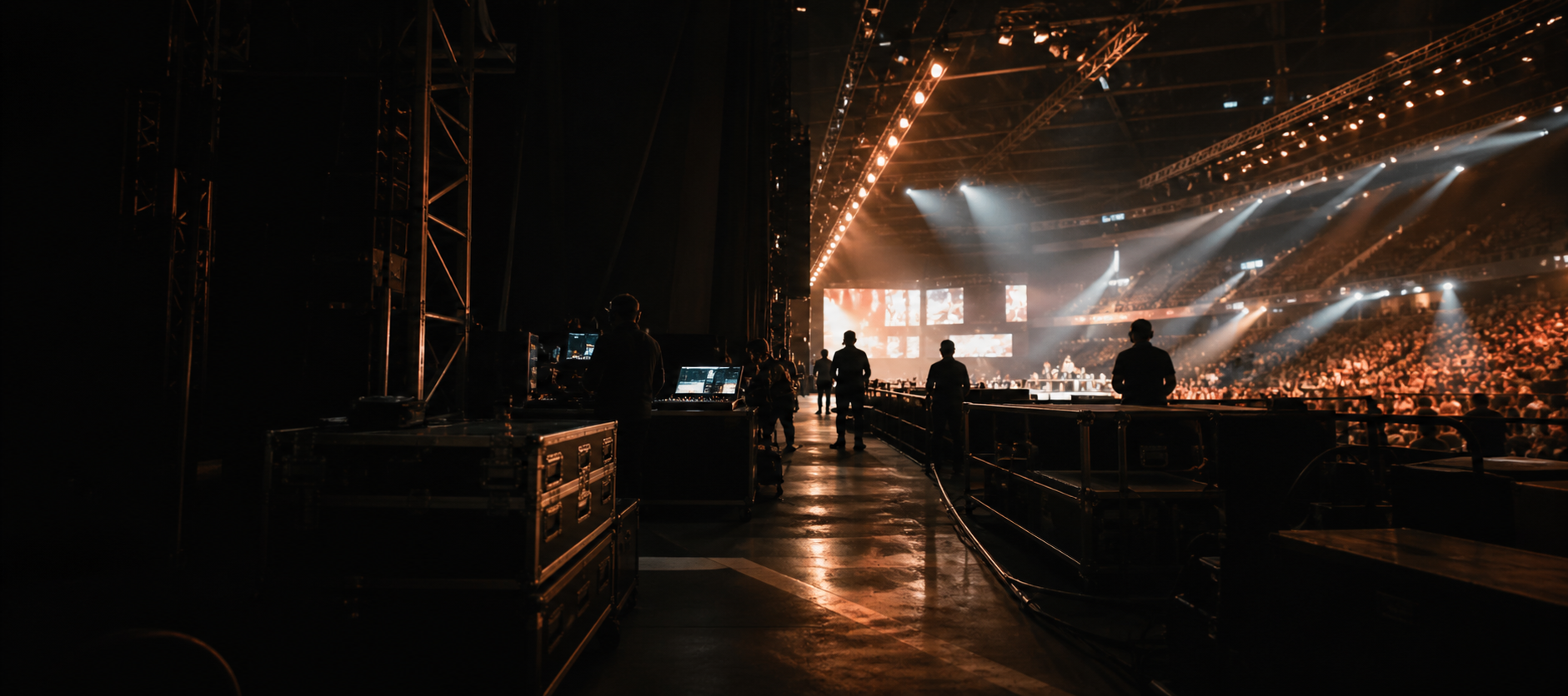 Backstage view of a live arena with production lights, crew silhouettes and a crowd in the distance.